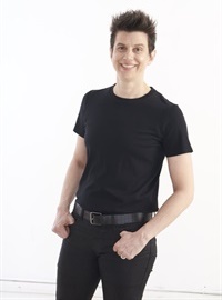 Instructor Jessica Hill, an athletic woman with short black hair, dressed in black, smiling and standing with her thumbs in her pockets